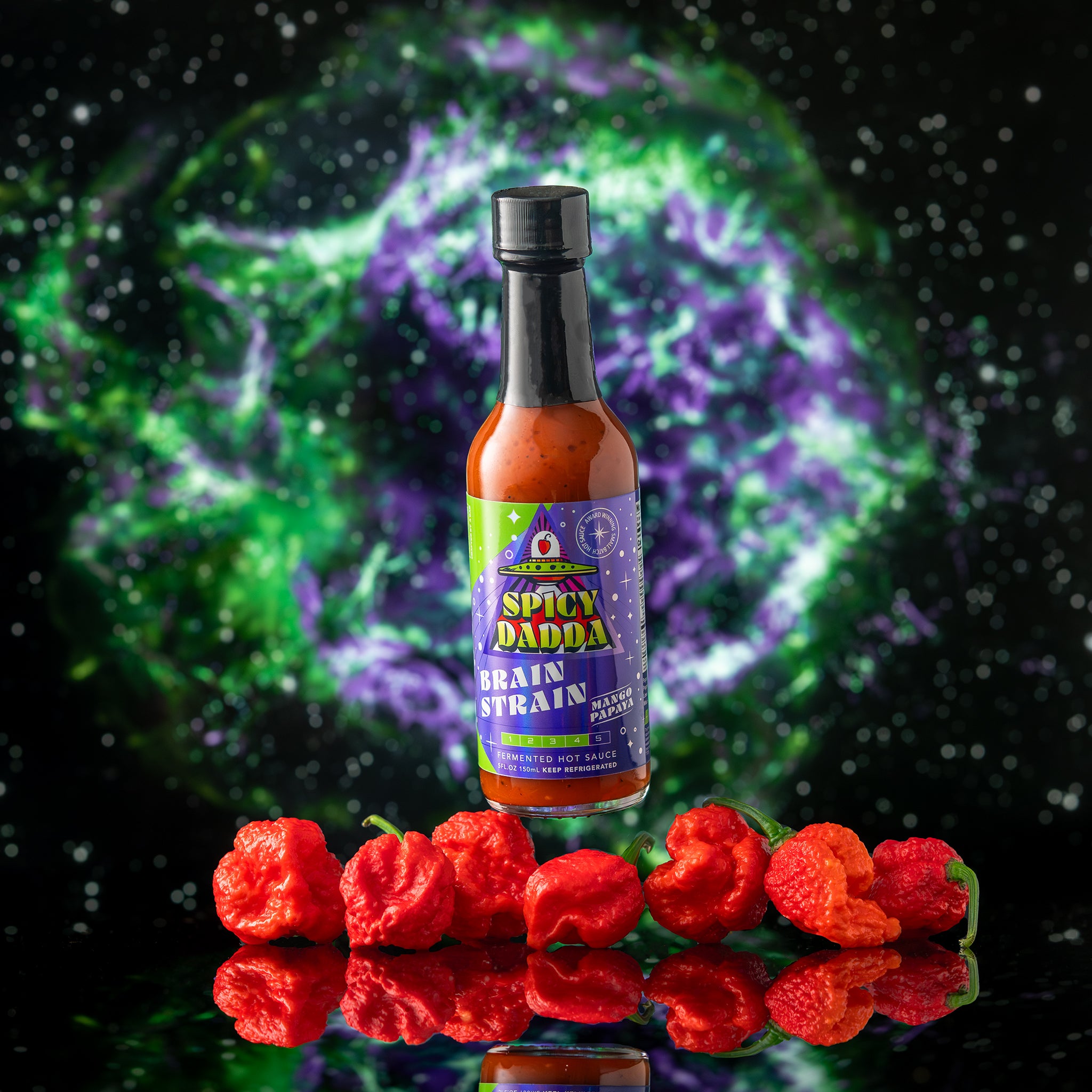 Bottle of Spicy Dadda Brain Strain Mango Papaya hot sauce surrounded by homegrown red superhot Brain Strain peppers against a glowing cosmic space background.