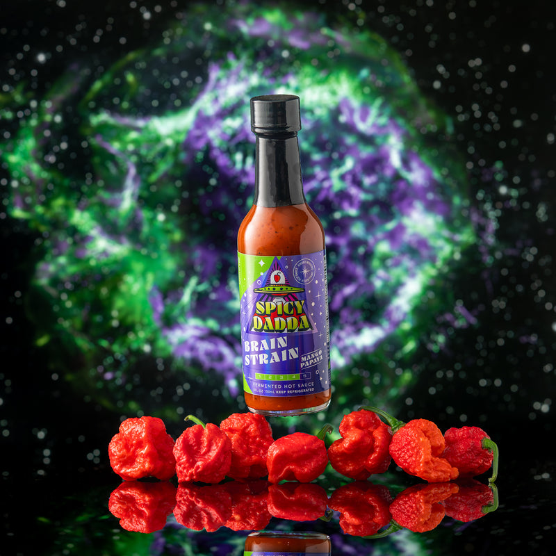 Bottle of Spicy Dadda Brain Strain Mango Papaya hot sauce surrounded by homegrown red superhot Brain Strain peppers against a glowing cosmic space background.